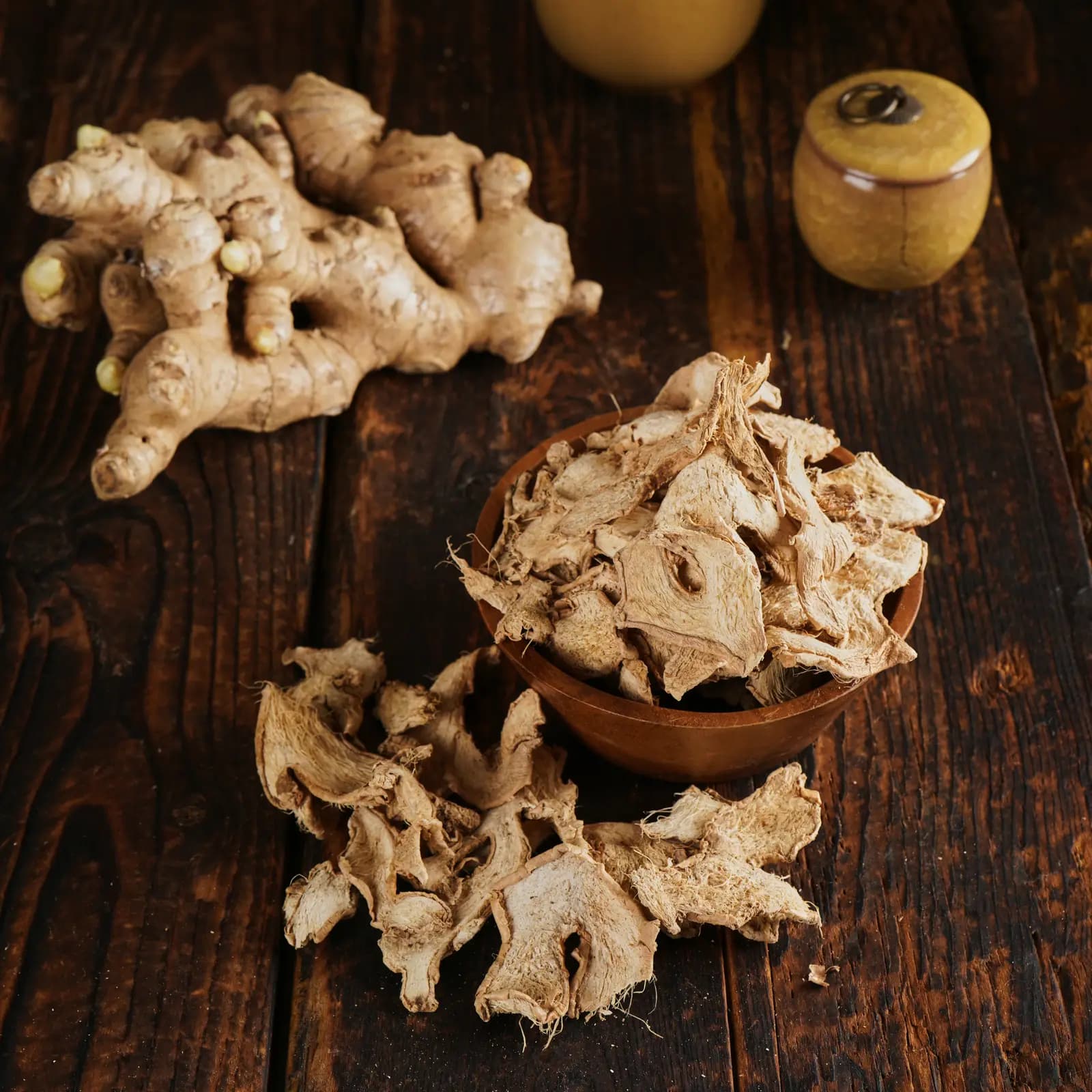Bowl of dried ginger slices beside a fresh Jahe Gajah rhizome on a wooden table